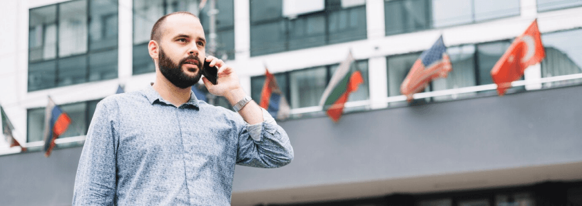 Profissional de tecnologia falando ao telefone em um ambiente urbano com bandeiras de diversos países ao fundo, representando conexões internacionais e oportunidades no mercado europeu de TI